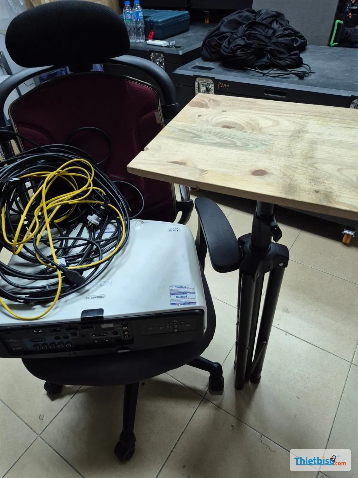 Máy chiếu và dây cáp trên ghế, cạnh bàn gỗ gắn chân đỡ và thùng thiết bị trong kho