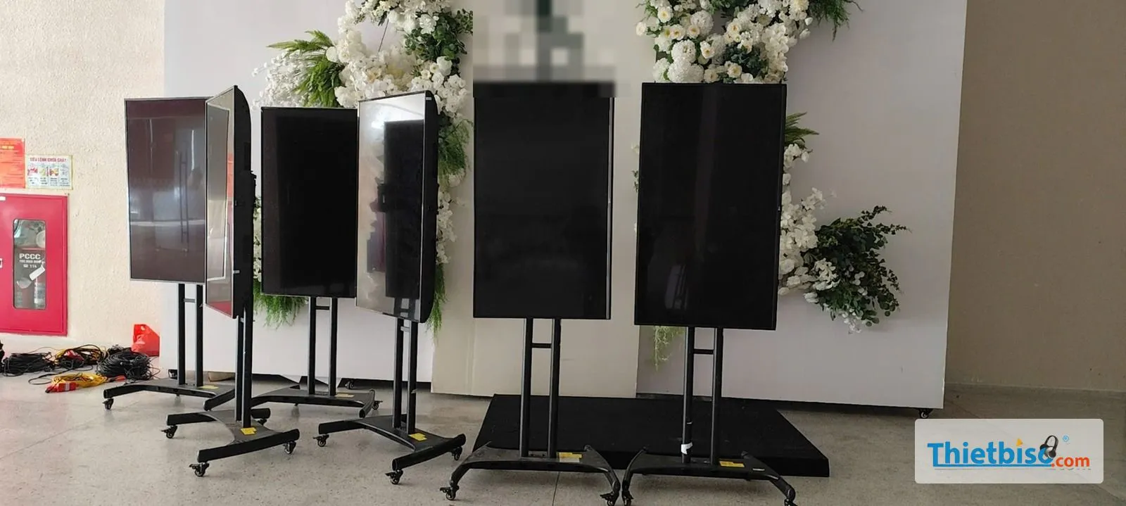 Năm tivi đặt dọc trên chân đế có bánh xe, xếp trước backdrop hoa trắng tại sảnh sự kiện