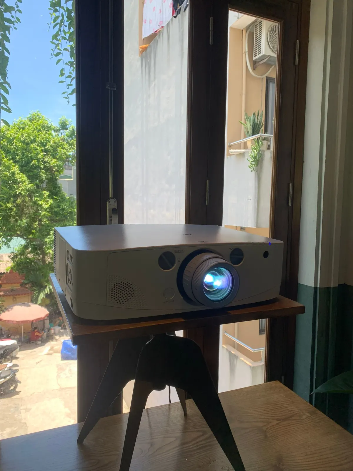 Máy chiếu cỡ lớn đặt trên giá ba chân cạnh cửa sổ, ống kính sáng xanh