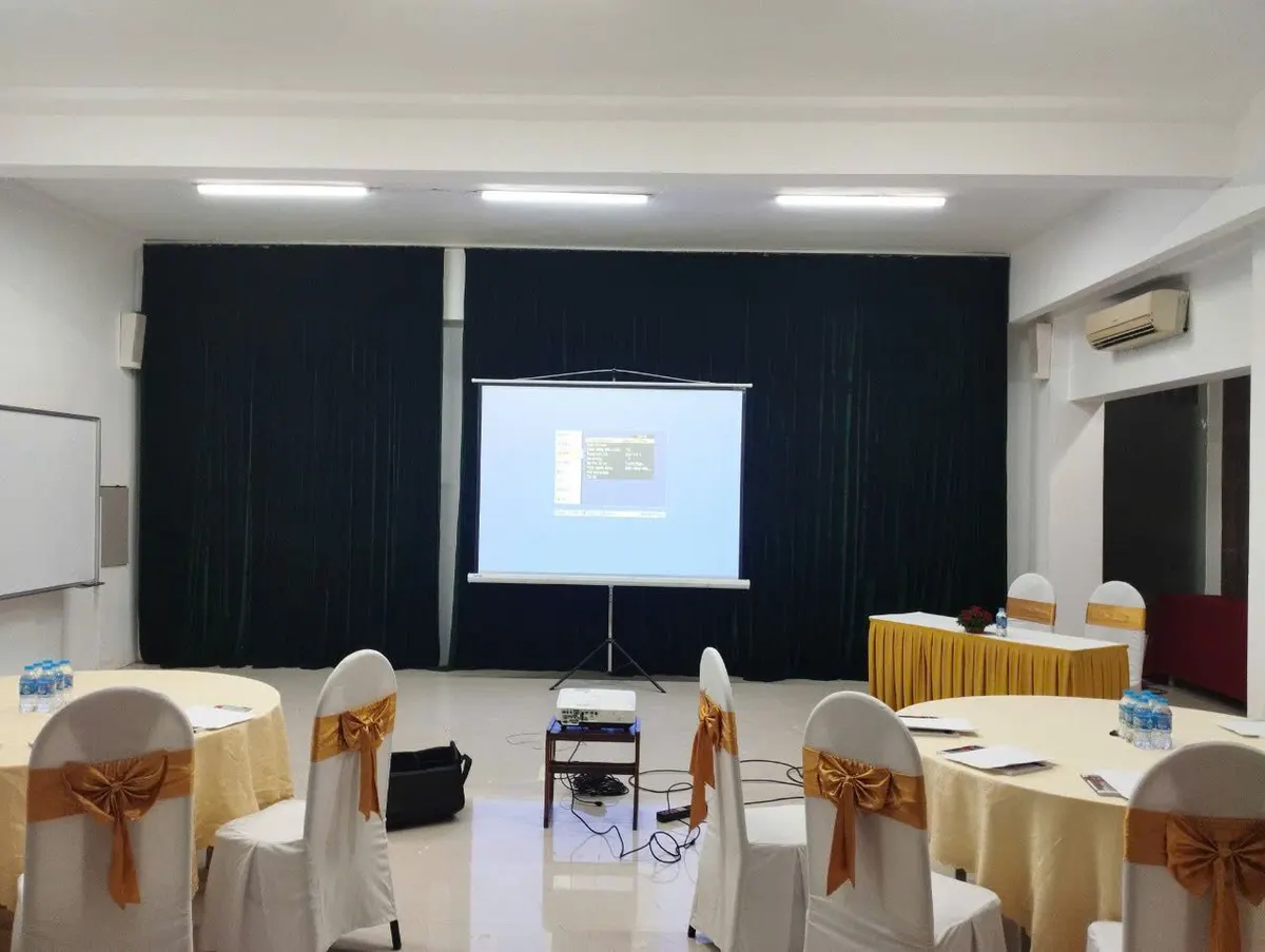 Phòng tiệc có bàn tròn ghế phủ nơ, màn chiếu chân đặt trước rèm đen